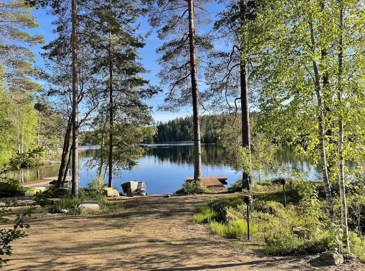 hiekkaranta, jossa on mäntyjä ja kasi laituria sekä vene, sininen taivas ja järvi