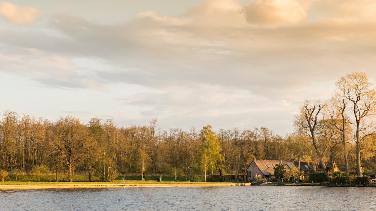Scenic view of a lakeside cottage surrounded by autumn trees under a warm sunset sky.